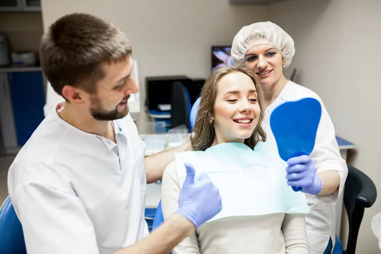 Une patiente sourit en découvrant son sourire dans un miroir, entourée du médecin-dentiste et de l’assistante